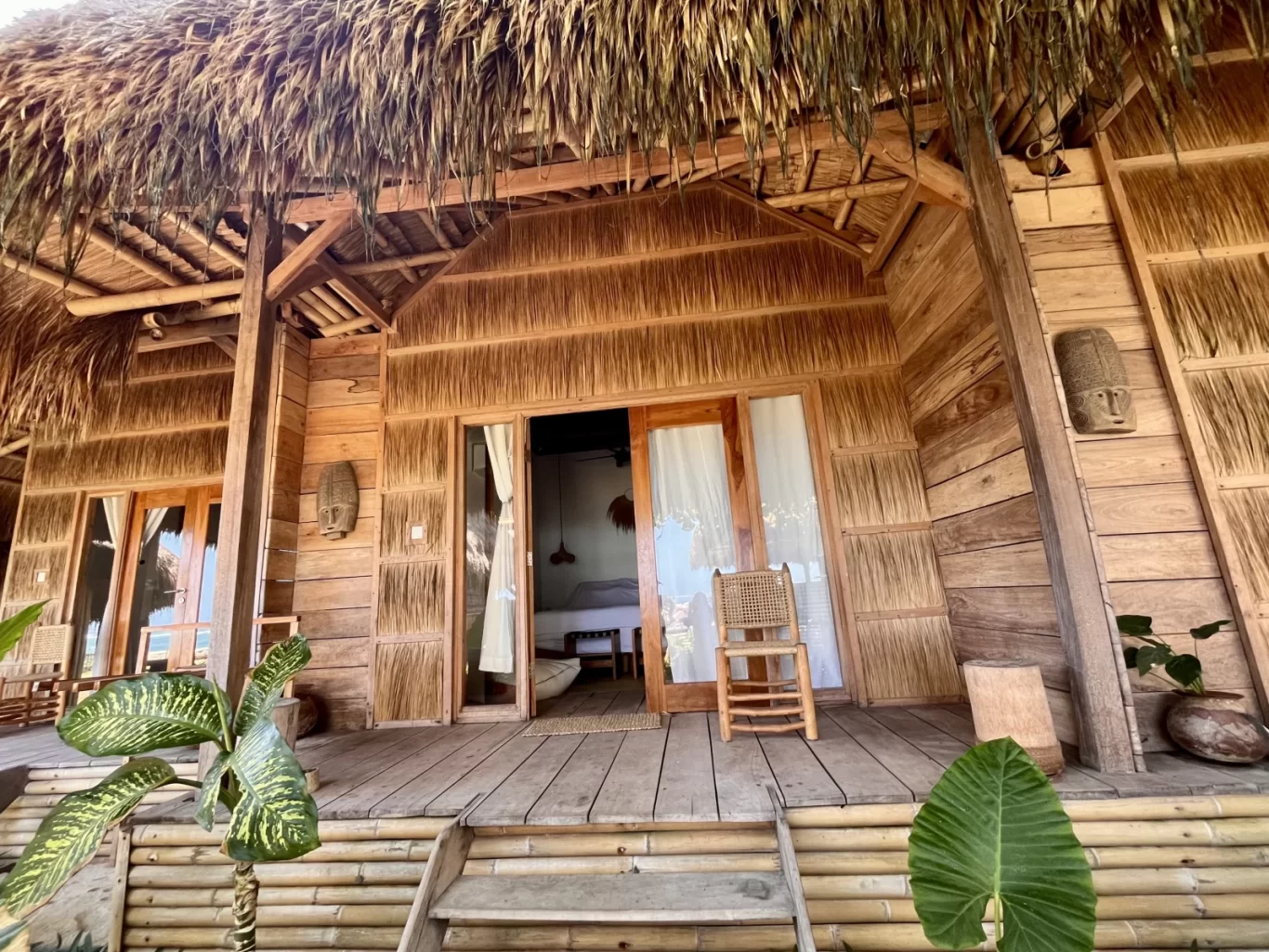A rustic wooden spa bungalow with a thatched roof and a bamboo porch features an open doorway revealing a bed, surrounded by green tropical plants.