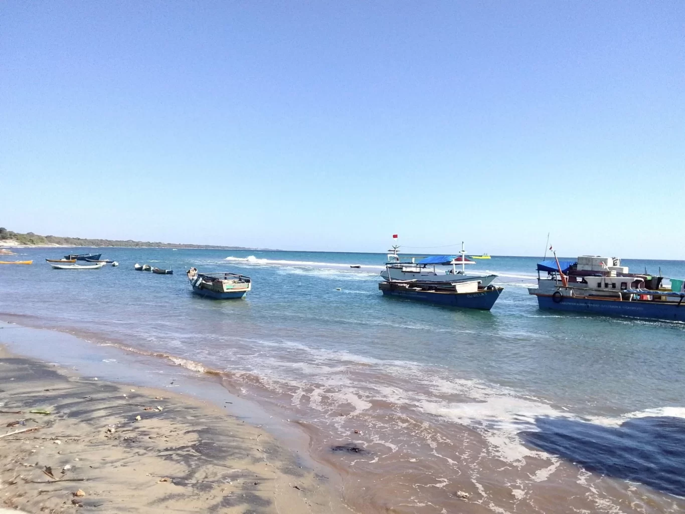 A sandy beach with dark wet sand meets clear blue-green ocean water where several blue boats are anchored under a bright clear sky.