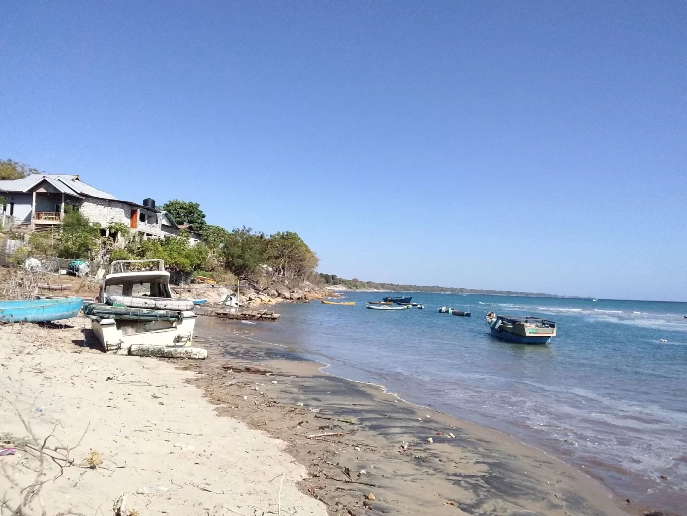 A sandy beach with several small boats, some on shore and others in the blue-green ocean, fronts houses on a green hillside under a clear blue sky.