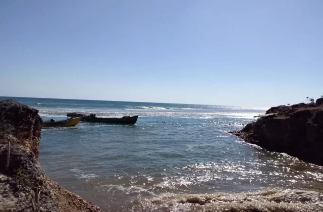 Two small boats, one yellow and one dark, float in blue-green ocean water between rocky cliffs under a clear blue sky. Sunlight sparkles on the water.