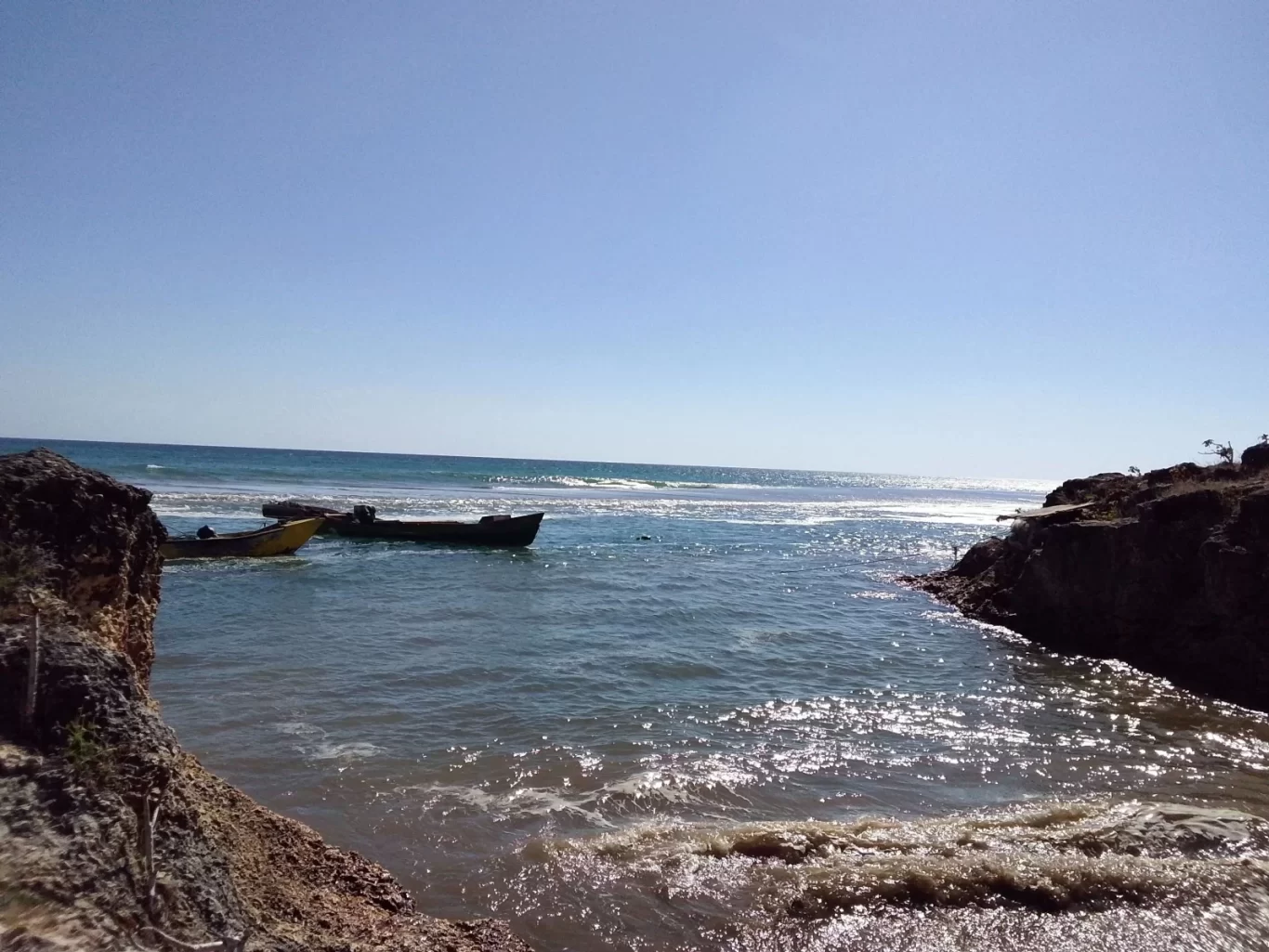 Two small boats, one yellow and one dark, float in blue-green ocean water between rocky cliffs under a clear blue sky. Sunlight sparkles on the water.
