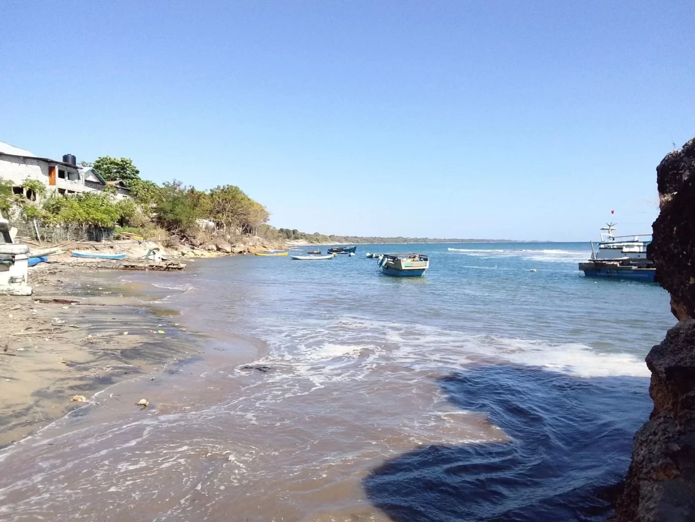 A coastal scene shows several small boats floating on blue-green water next to a sandy beach with buildings and trees on the shore, under a clear blue sky.
