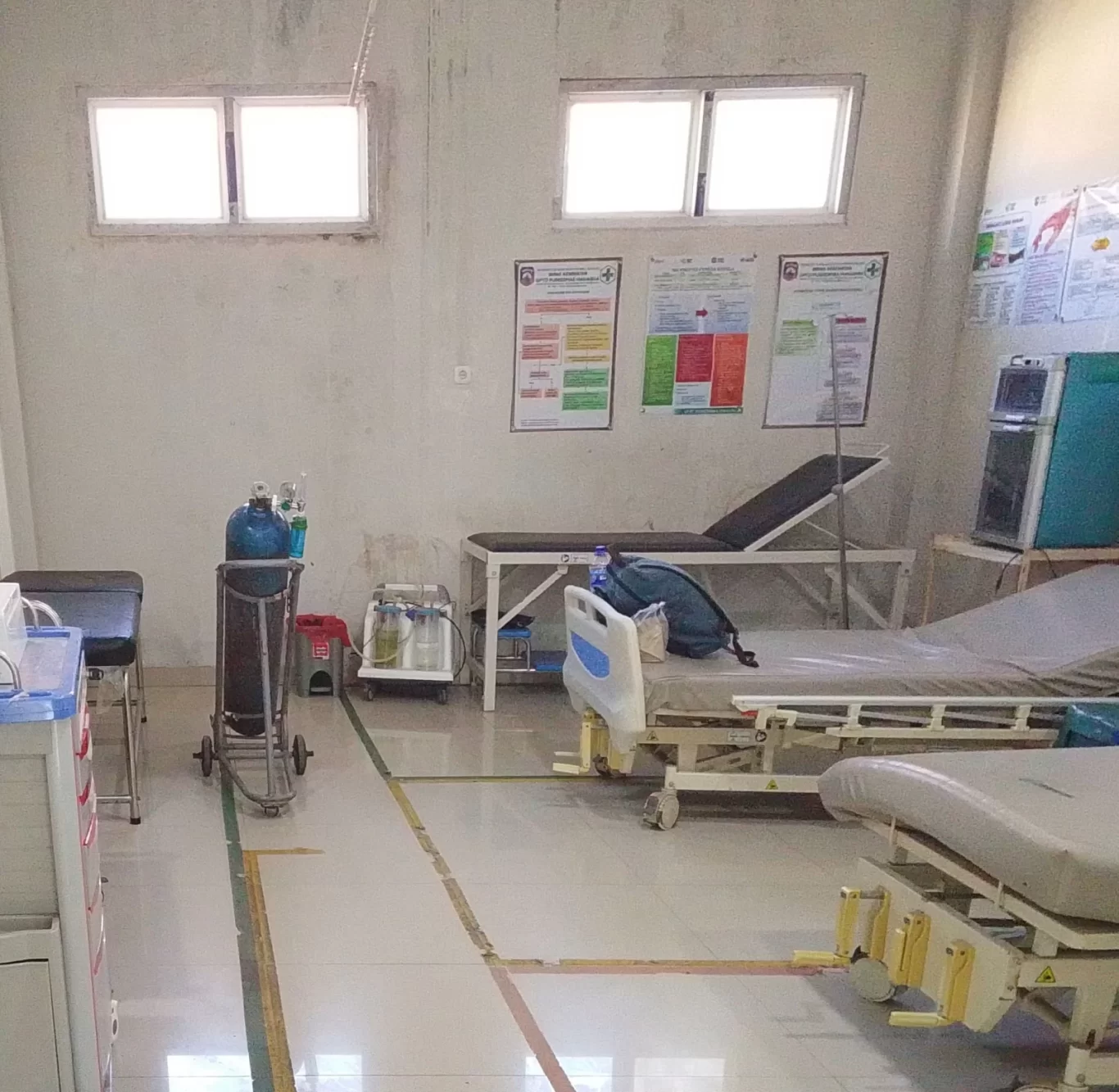 A sparsely furnished medical room features two hospital beds, two examination tables, an oxygen tank, and several health posters on light grey walls with two windows.