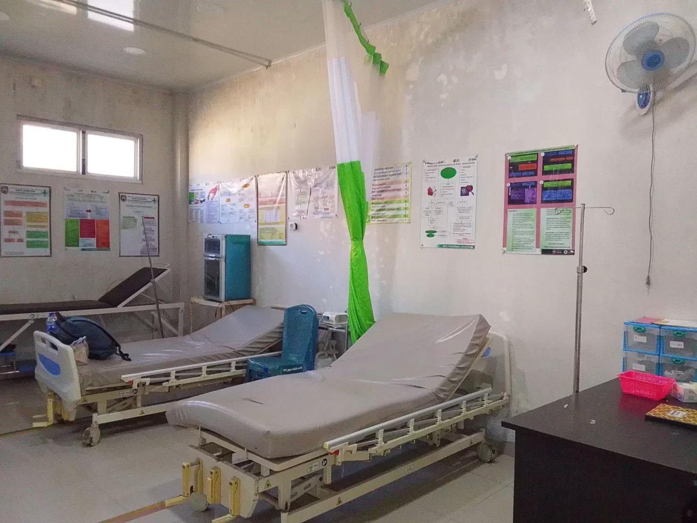 A medical room with three empty hospital beds, white walls adorned with posters, a green and white privacy curtain, and a wall fan.