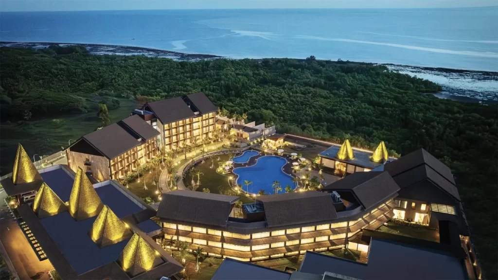 An aerial view shows a luxurious coastal resort illuminated at dusk, featuring multiple buildings, a large blue swimming pool, and lush green trees bordering the dark blue ocean.
