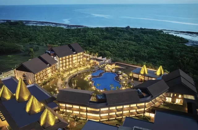 An aerial view shows a luxurious coastal resort illuminated at dusk, featuring multiple buildings, a large blue swimming pool, and lush green trees bordering the dark blue ocean.
