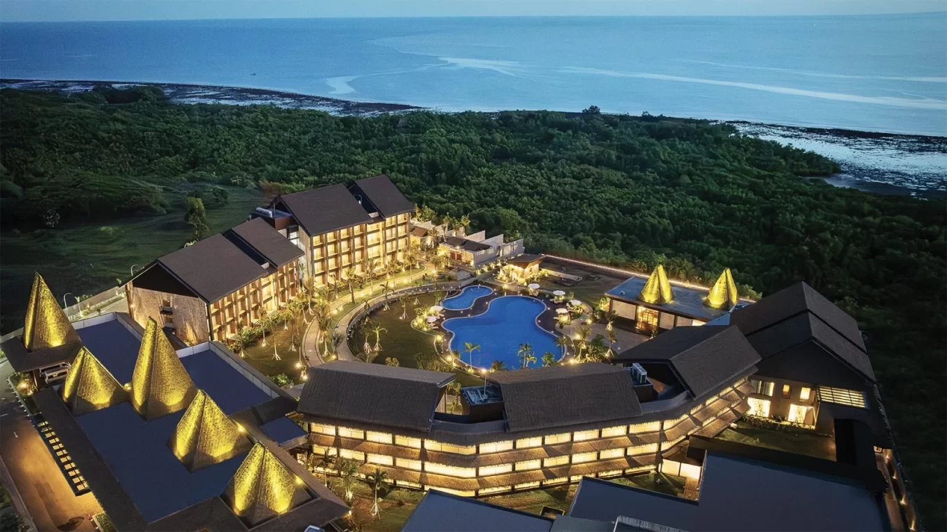 An aerial view shows a luxurious coastal resort illuminated at dusk, featuring multiple buildings, a large blue swimming pool, and lush green trees bordering the dark blue ocean.
