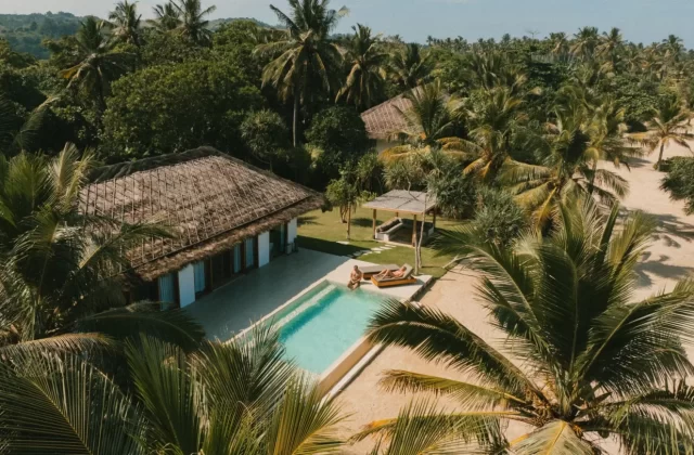 An aerial view shows a thatched-roof villa with a swimming pool, where two people relax, surrounded by dense green palm trees and sandy ground under a clear sky.