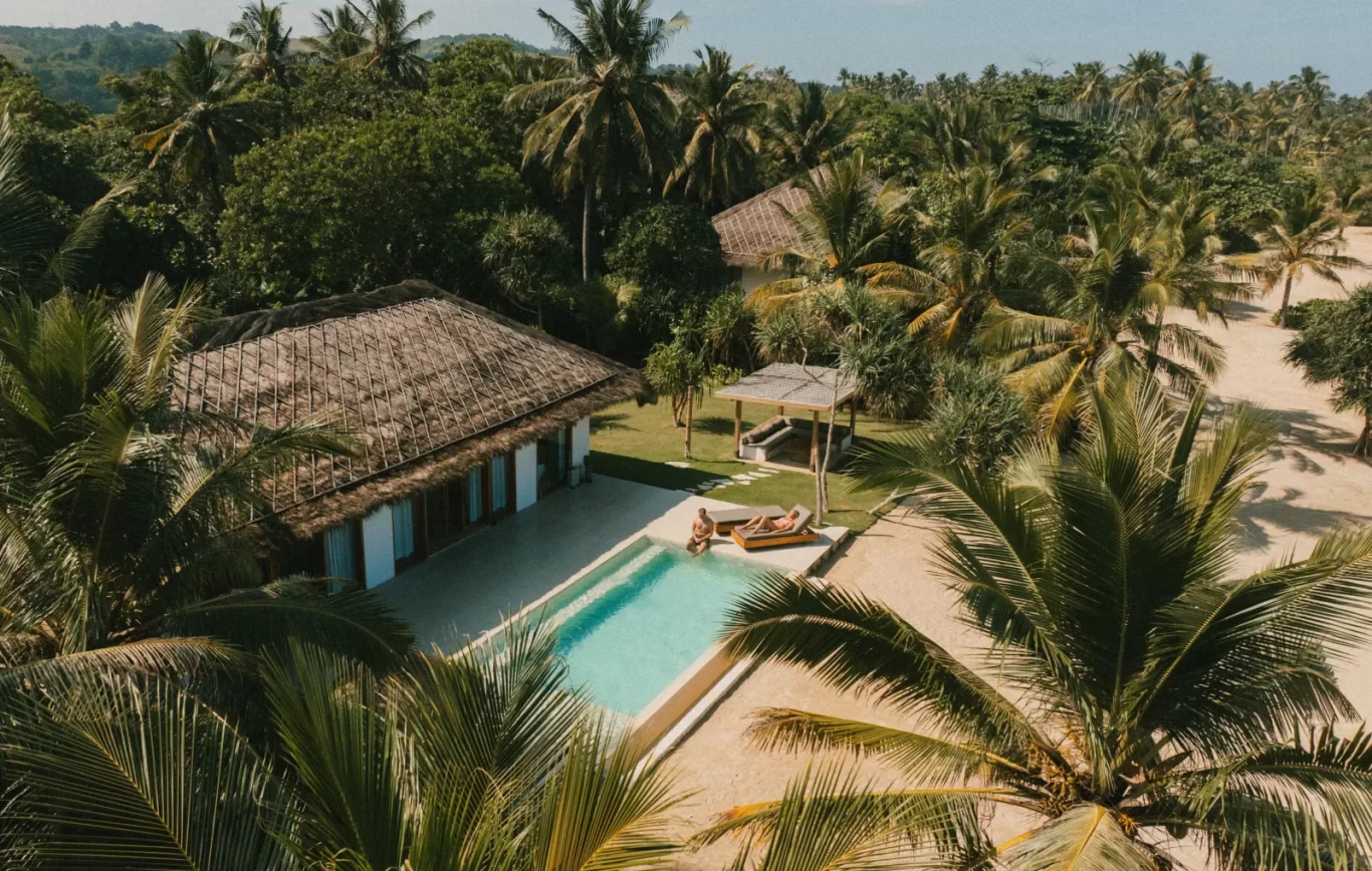 An aerial view shows a thatched-roof villa with a swimming pool, where two people relax, surrounded by dense green palm trees and sandy ground under a clear sky.