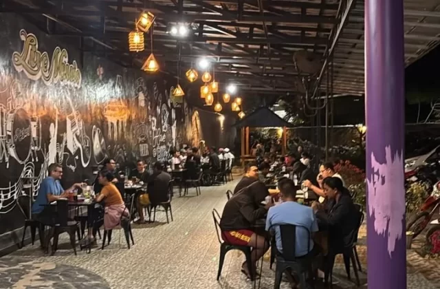 A lively outdoor restaurant at night, with patrons seated at tables under a metal roof, illuminated by warm hanging lights. A black wall features white 'Live Music' graffiti and musical instrument art.