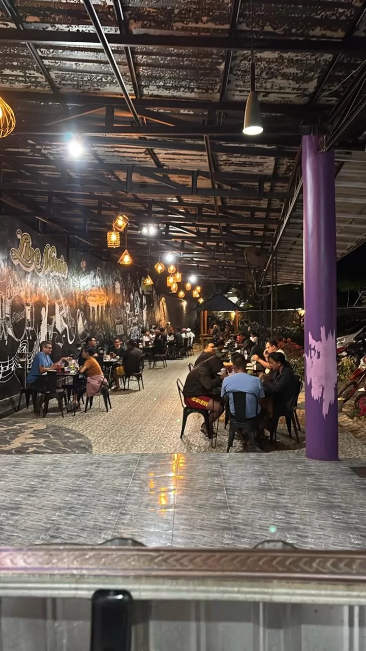 A lively outdoor restaurant at night, with patrons seated at tables under a metal roof, illuminated by warm hanging lights. A black wall features white 'Live Music' graffiti and musical instrument art.