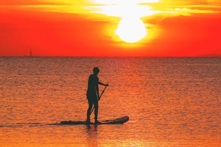 A person in silhouette stands on a paddleboard in calm, orange-hued water, facing a brilliant orange and yellow sunset with a bright white sun.
