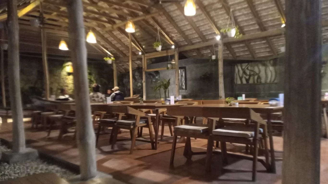 A dimly lit, open-air restaurant features a high thatched roof, wooden tables and chairs, and warm pendant lights. Several blurry patrons are seated, and hanging plants adorn the space.