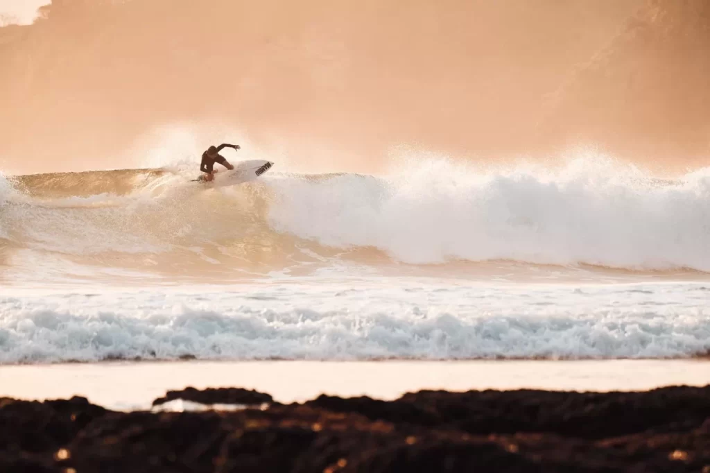 A surfer in a dark wetsuit rides a large, breaking wave in the ocean, with a golden-orange sky and dark rocks in the foreground.