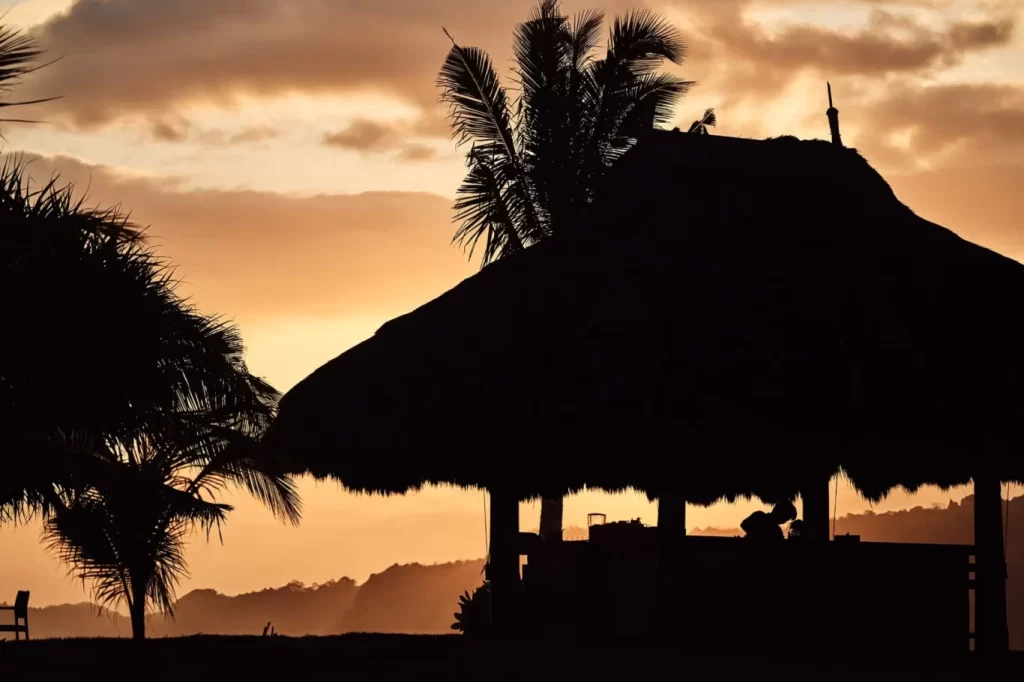 A large thatched-roof hut and palm trees are silhouetted against a warm orange and yellow sunset sky with clouds over distant hills.