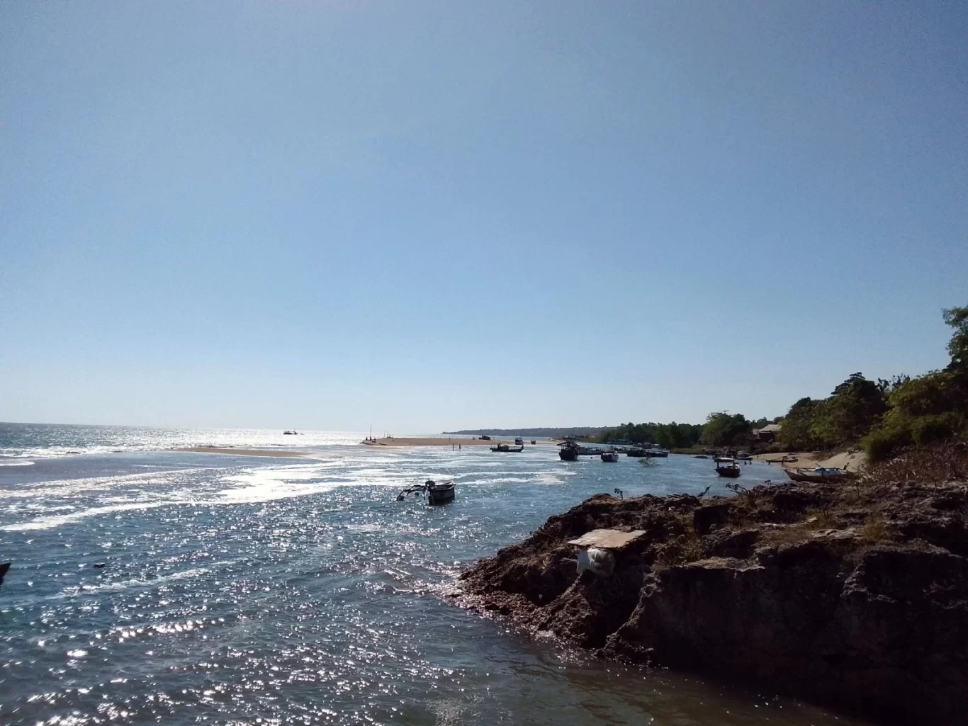 A sunny coastal scene shows sparkling blue water with numerous small fishing boats, a rocky shore on the right, and a distant sandy beach under a clear blue sky.