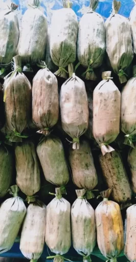 Many cylindrical food items wrapped in dried banana leaves and tied with natural string are arranged closely in rows, displaying muted green, brown, and off-white tones.