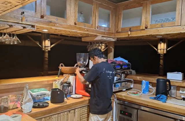 A person in a black shirt and light shorts operates a red espresso machine in a rustic kitchen with woven bamboo ceilings and wooden counters.