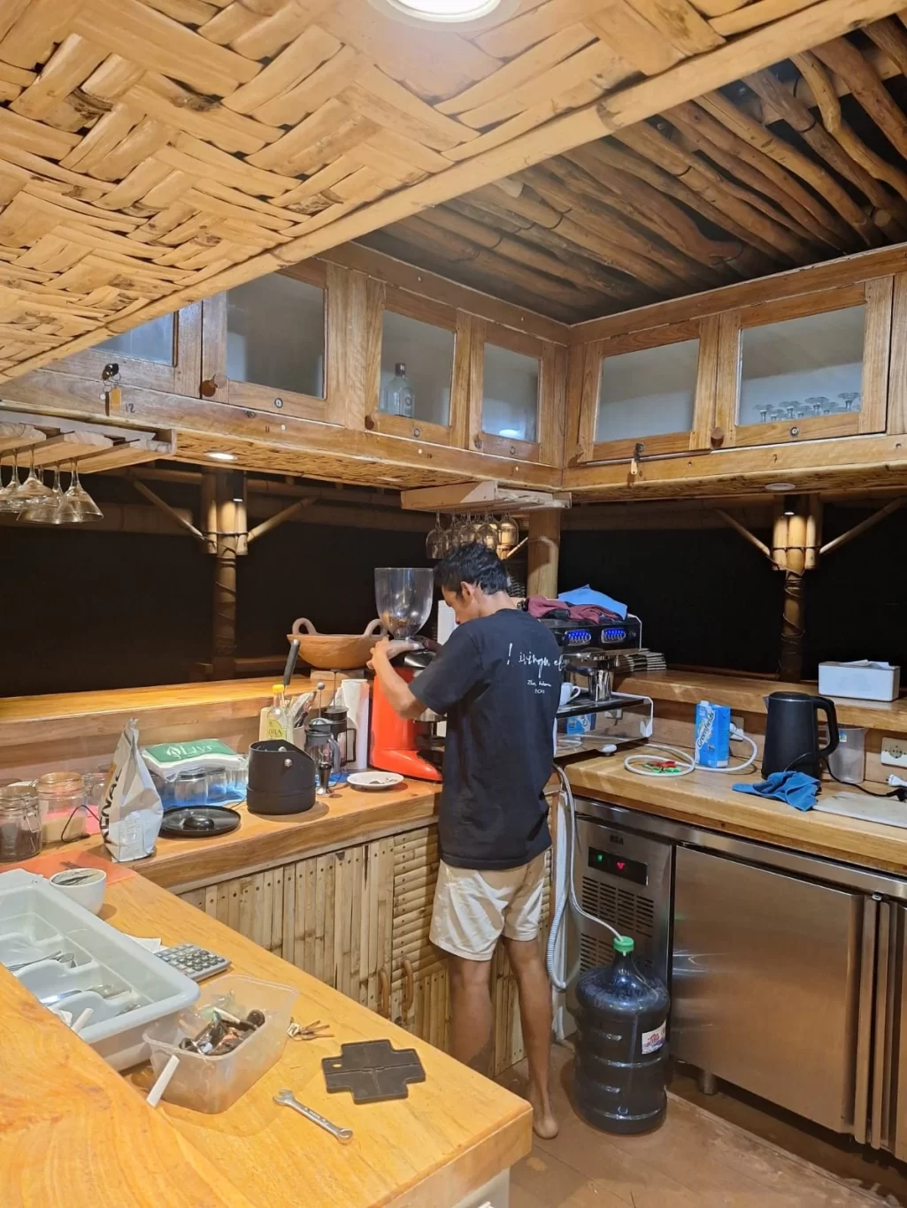 A person in a black shirt and light shorts operates a red espresso machine in a rustic kitchen with woven bamboo ceilings and wooden counters.