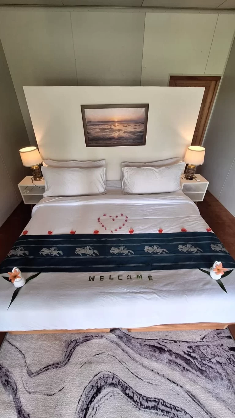 A neatly made hotel bed with white linens, a dark blue patterned runner, and pink rose petals forming a heart, flanked by two lamps and a sunset painting.