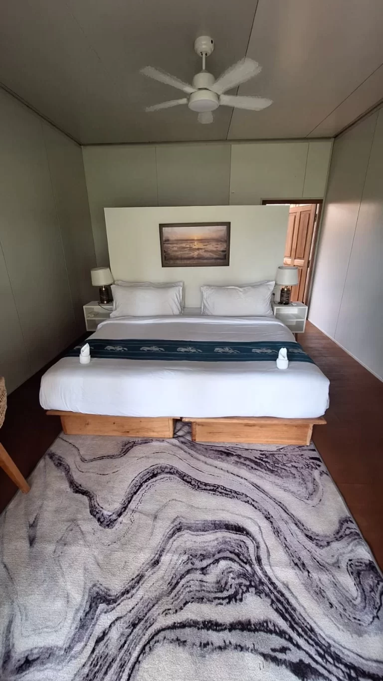 A resort bedroom features a large bed with white linens, a blue runner, a grey marbled rug, light walls, a ceiling fan, and a framed landscape.