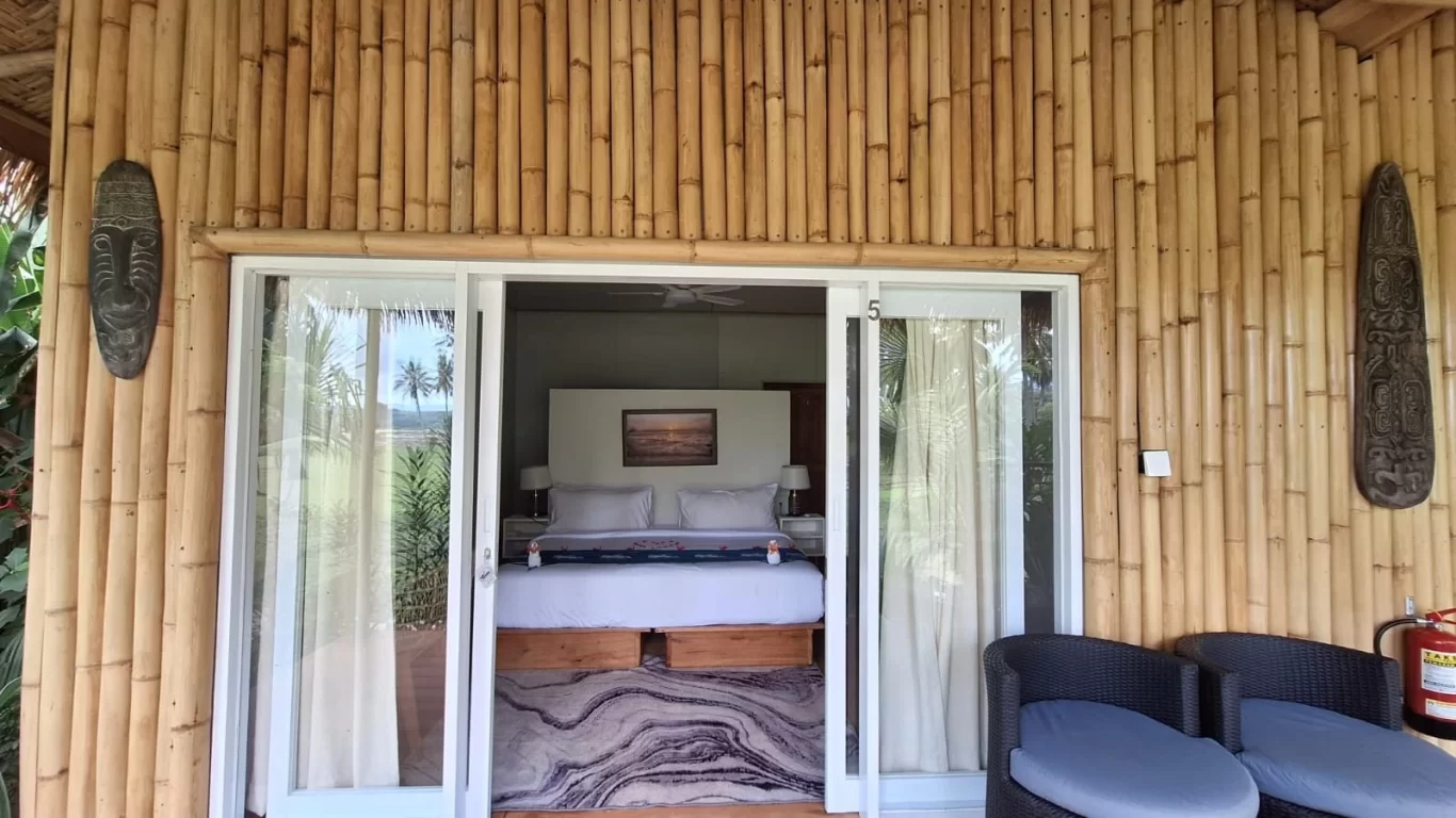 A bamboo-walled room entrance features a sliding glass door revealing a white-linened bed, with two wicker chairs outside. Palm trees and a lagoon reflect in the glass.