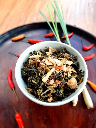 A white bowl of dark green leafy vegetables topped with crispy golden brown fried shallots sits on a dark wooden tray, surrounded by red chilies and green lemongrass.