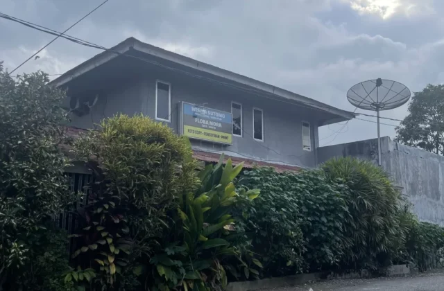 A grey two-story building, Wisma Sutomo, with a blue sign and a satellite dish, stands beside a paved road with green plants under a cloudy sky.