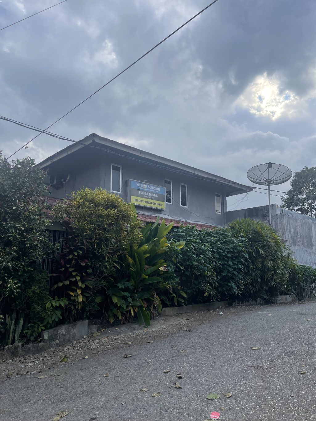 A grey two-story building, Wisma Sutomo, with a blue sign and a satellite dish, stands beside a paved road with green plants under a cloudy sky.