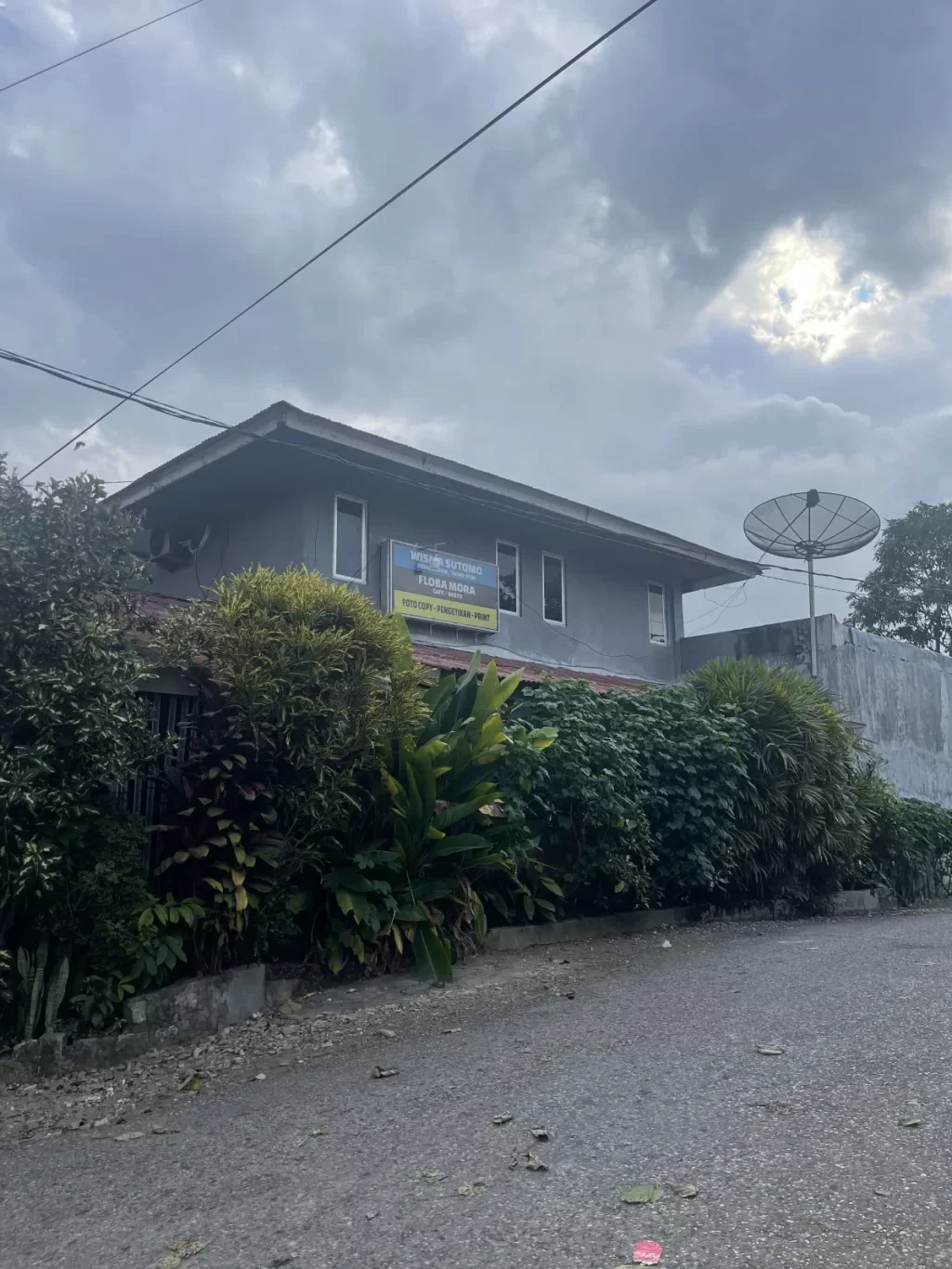 A grey two-story building, Wisma Sutomo, with a blue sign and a satellite dish, stands beside a paved road with green plants under a cloudy sky.