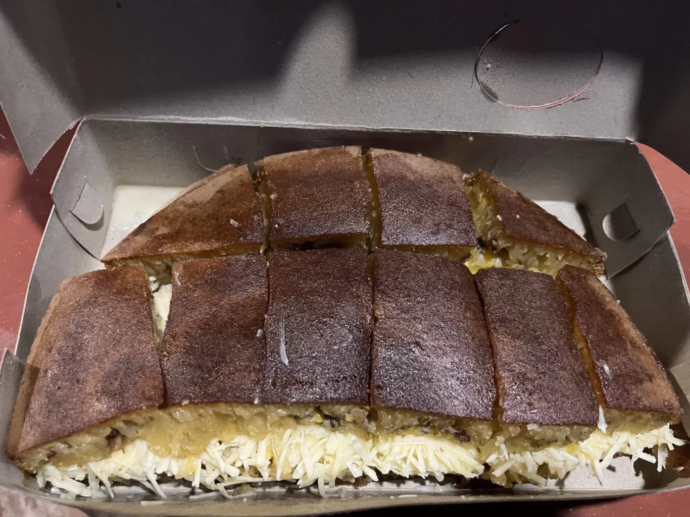 A sweet Indonesian pancake, Terang Bulan, is sliced into squares in a cardboard box, revealing a golden interior with shredded white cheese and condensed milk.