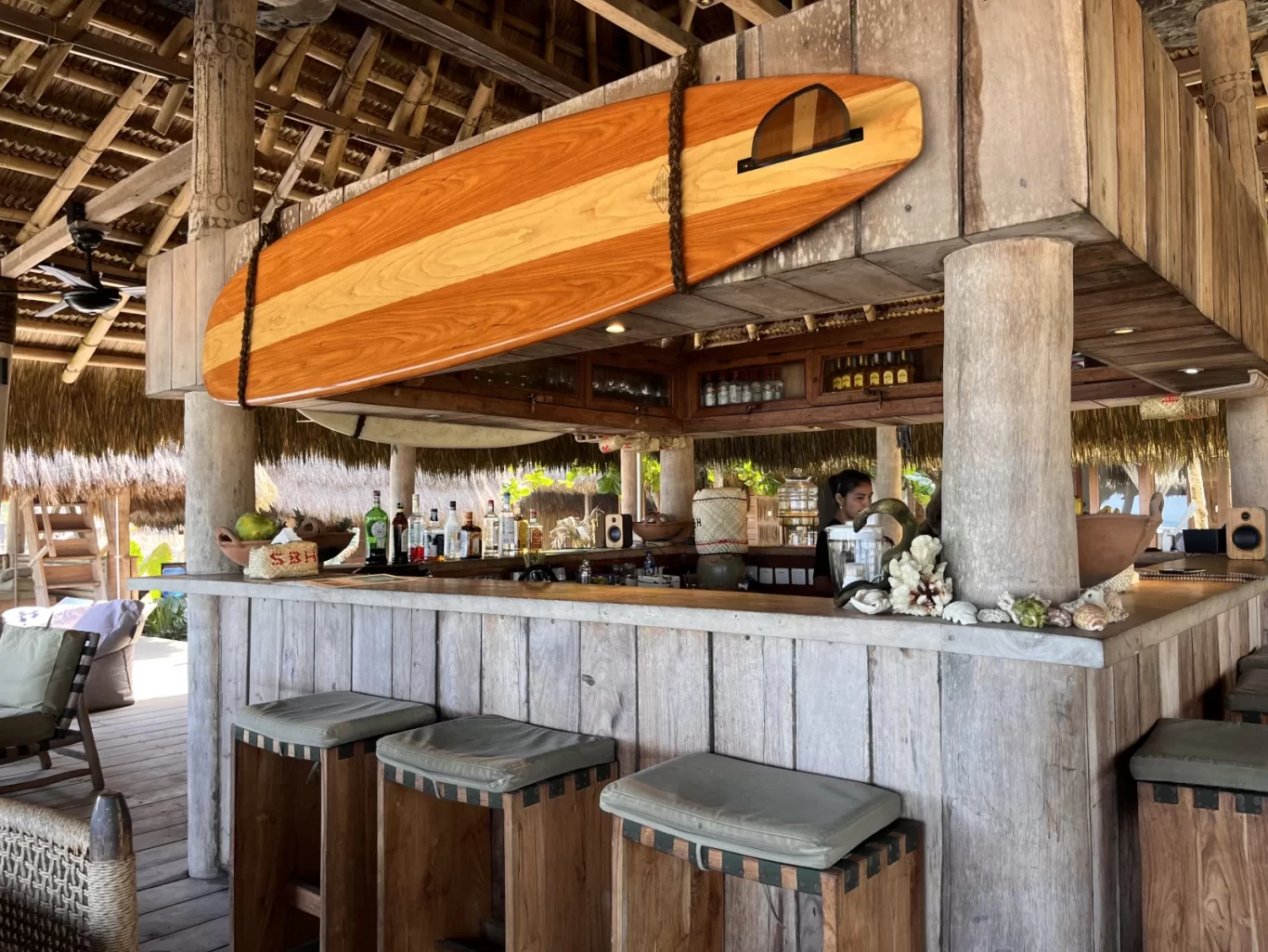 A rustic, open-air beach bar features a wooden counter with stools, a thatched roof, and a large wooden surfboard hanging above, with a bartender visible behind bottles.