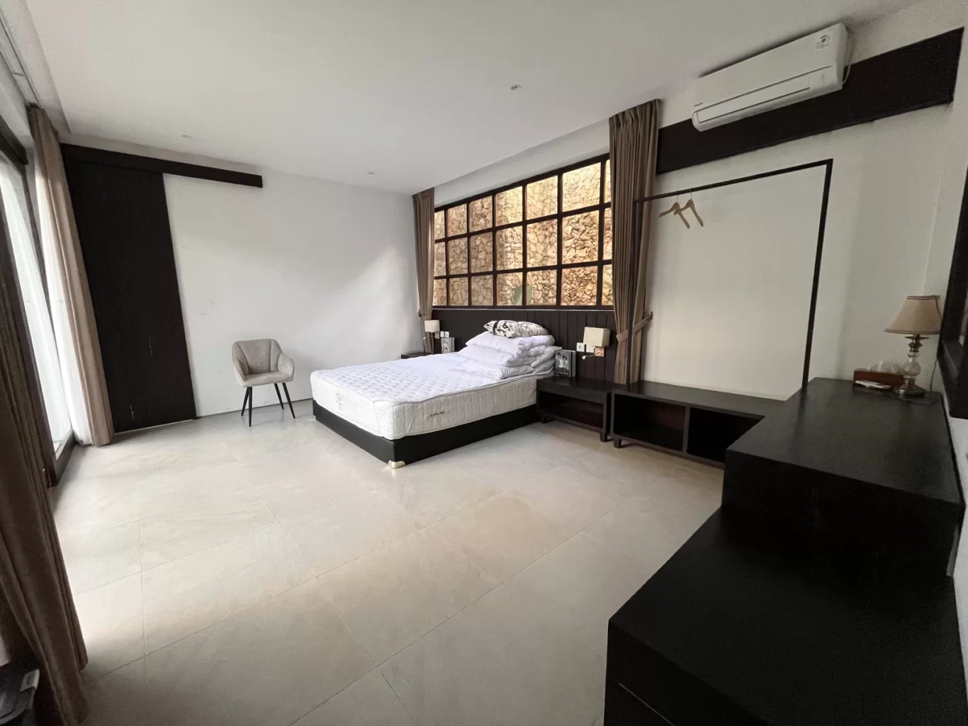A spacious modern bedroom features a white bed, dark wood furniture, light tiled floors, and a decorative window, with an air conditioner mounted above.