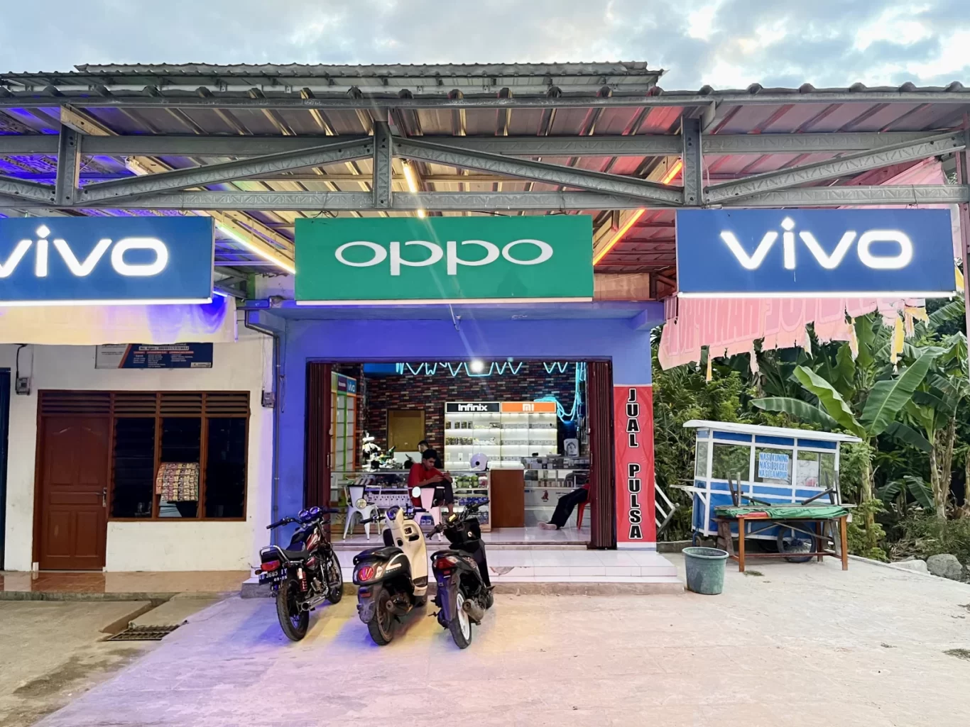 A blue-fronted phone repair shop, Boss Sparepart HP, in Tambolaka, Sumba, features glowing Vivo and Oppo signs, with three motorbikes parked outside under a cloudy sky.