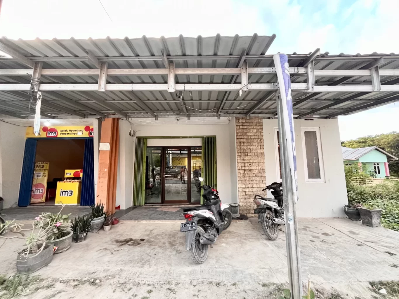 The exterior of Elsye Beauty Salon and an adjacent shop in Loura, Southwest Sumba, features light walls, green doors, and two parked motorbikes under a metal roof.