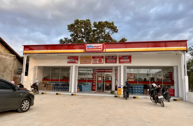 The red and white Alfamart minimarket in Tambolaka stands under a cloudy sky, with several motorcycles and a dark car parked outside.