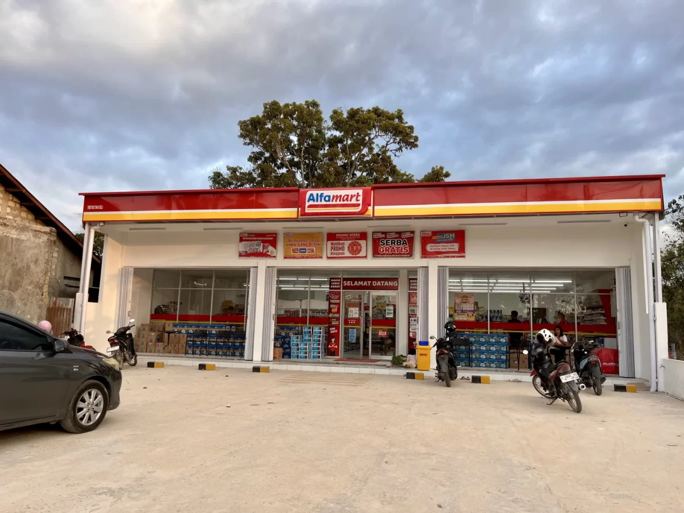 The red and white Alfamart minimarket in Tambolaka stands under a cloudy sky, with several motorcycles and a dark car parked outside.