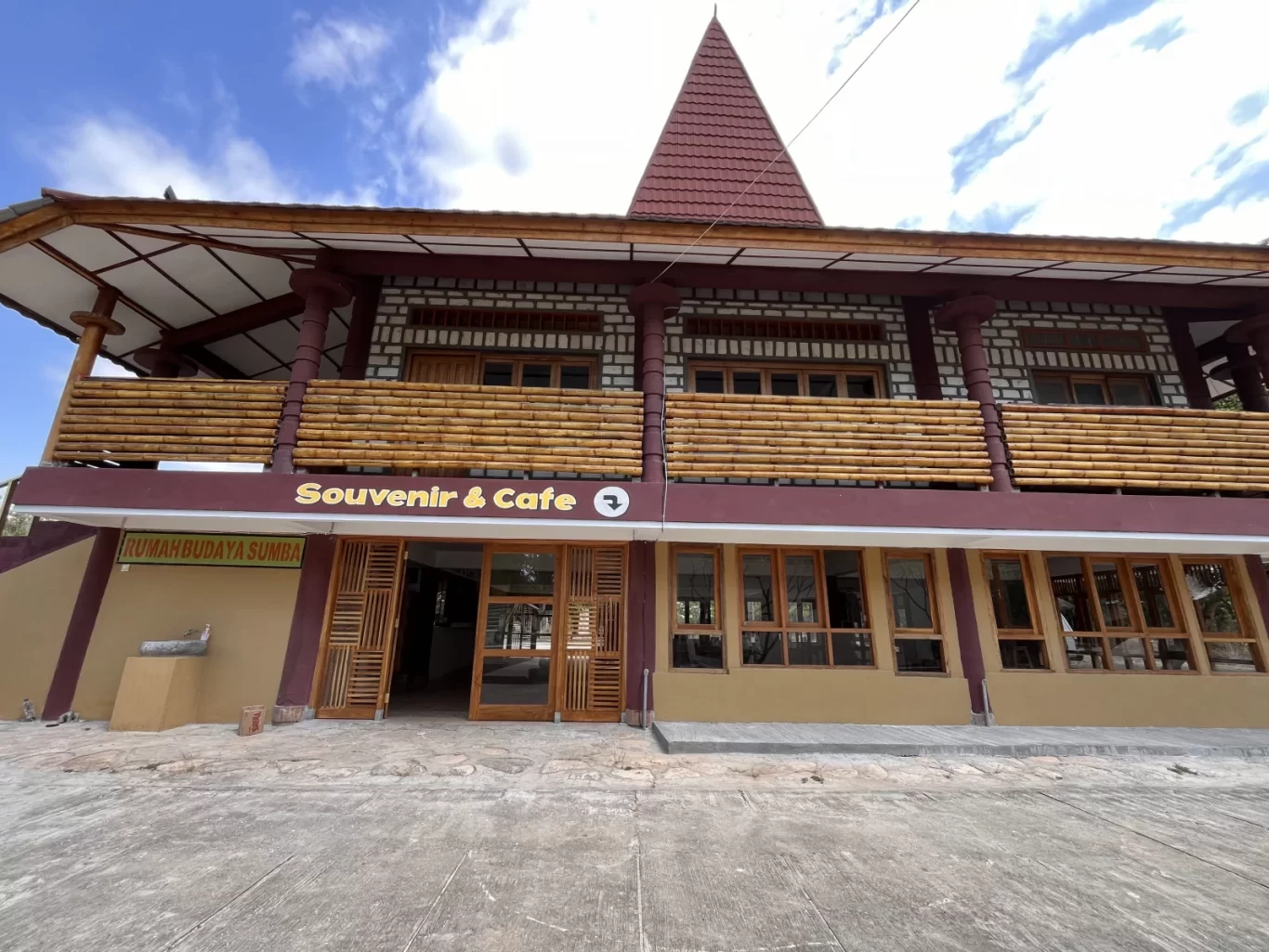 A two-story building with a red pointed roof, dark red and light brown walls, and a bamboo-railed balcony stands under a blue sky. The ground floor entrance has "Souvenir & Cafe" signage.