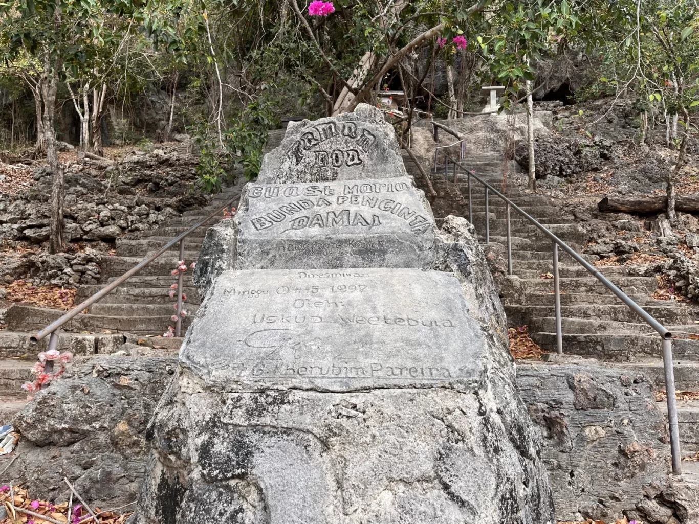 A large grey stone monument with carved text stands at the base of a long stone staircase ascending a rocky, tree-covered hillside.