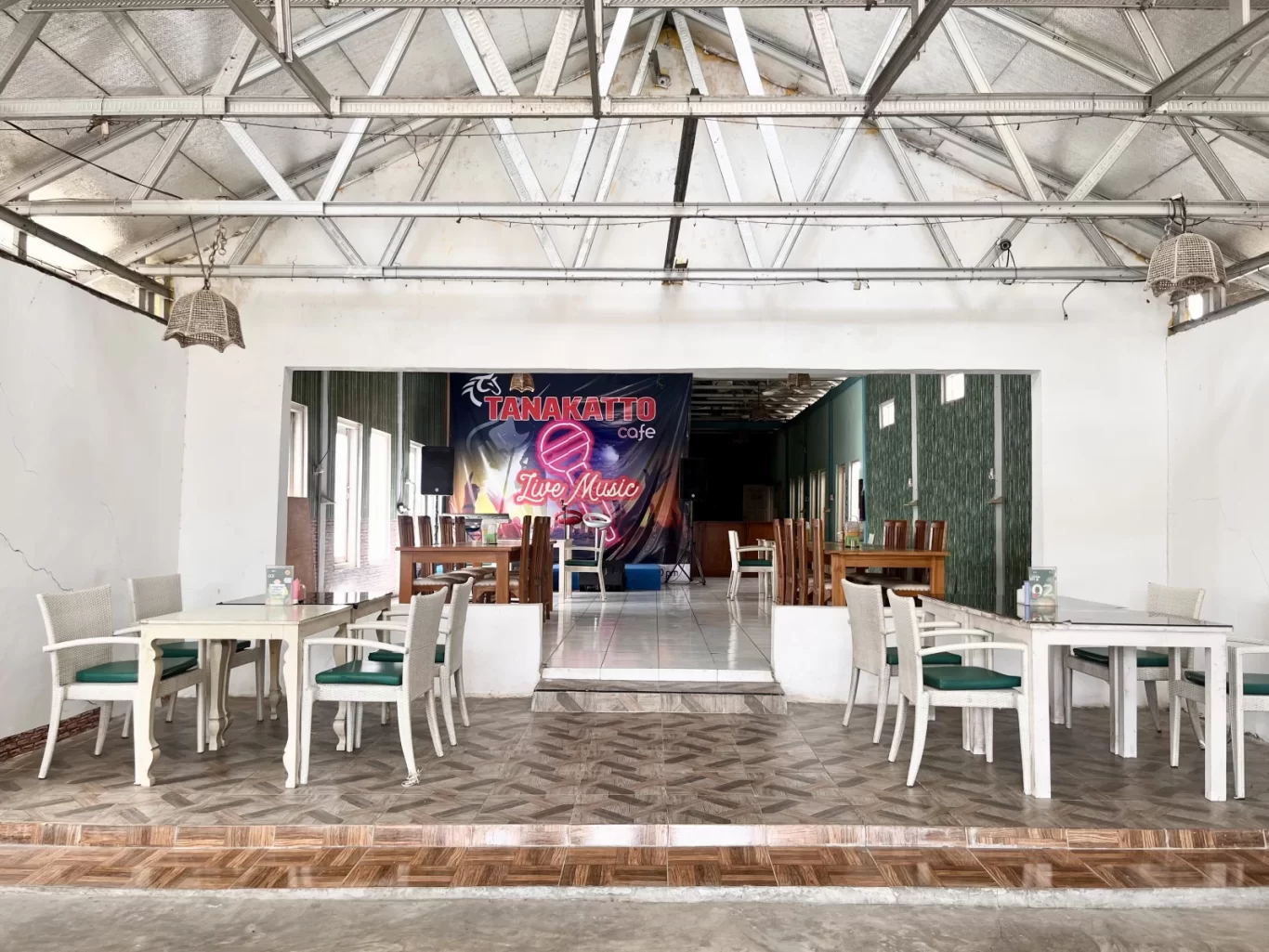 The bright, spacious interior of Tanakatto Restaurant in Waikabubak features white tables, green chairs, and a 'Live Music' banner.