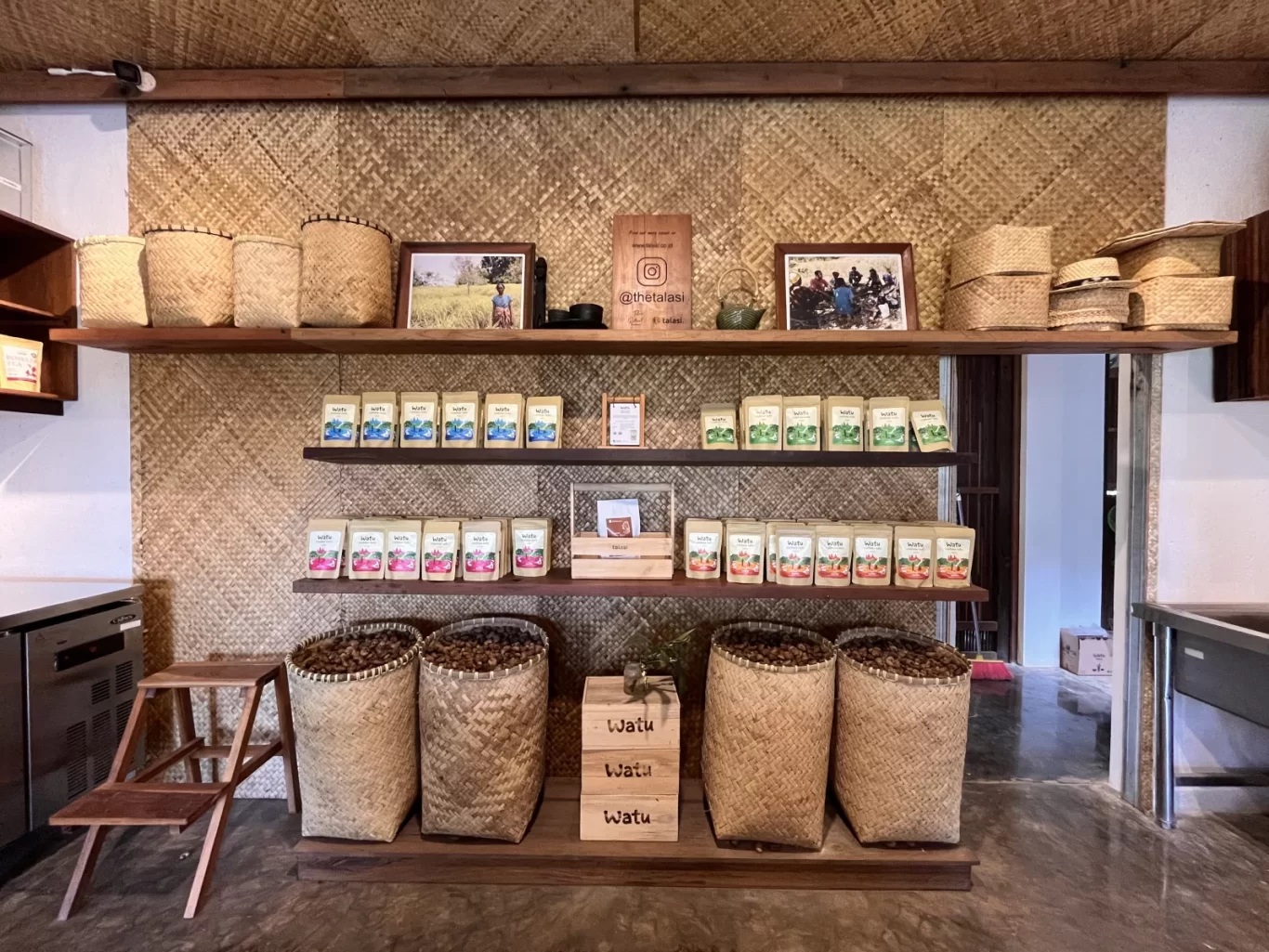 A coffee shop display wall features three wooden shelves holding woven baskets, framed photos, colorful coffee pouches, and large baskets of coffee beans against a woven straw background.