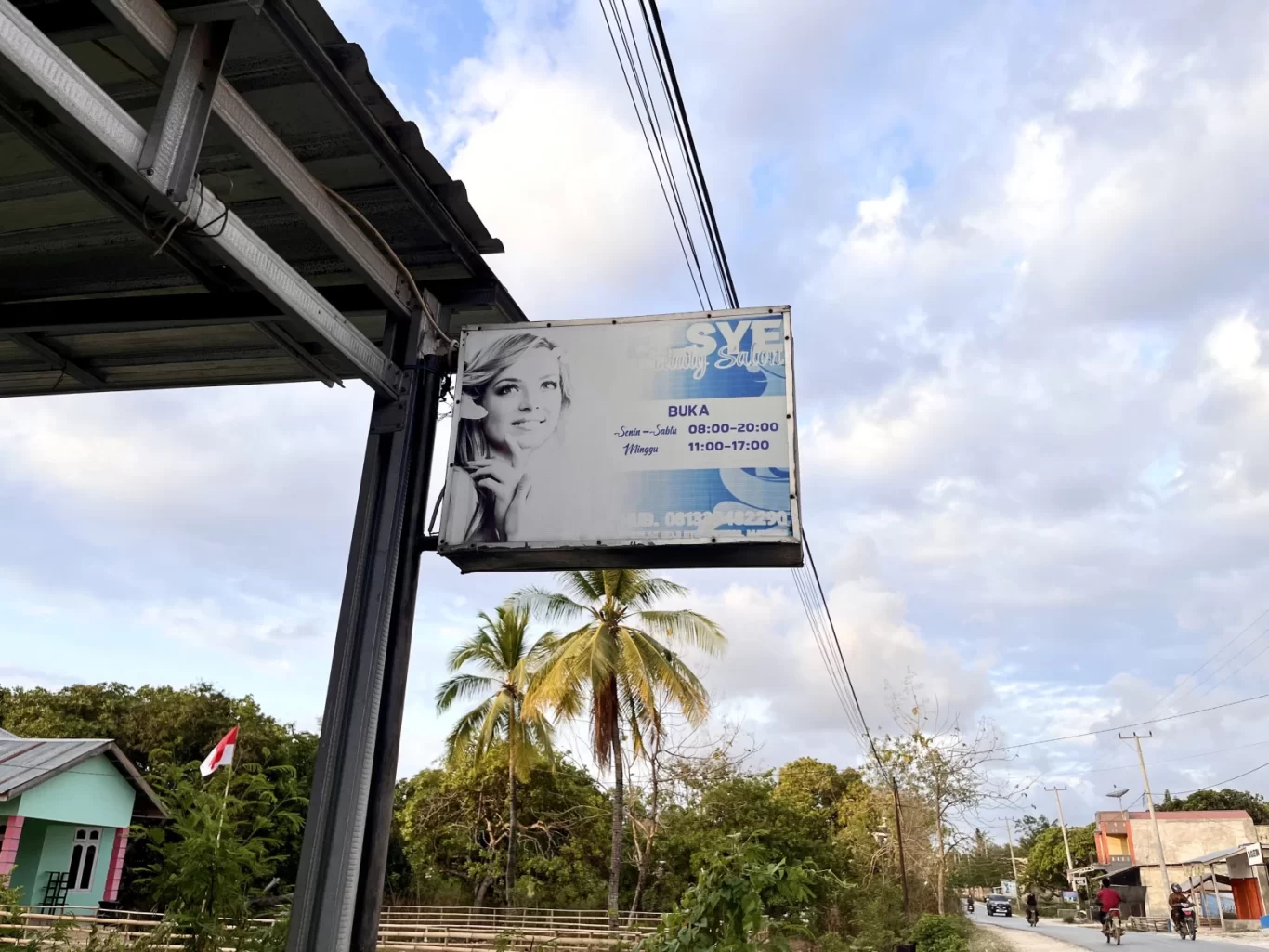 A white Elsye Beauty Salon sign featuring a woman's face and operating hours hangs from a metal structure against a partly cloudy sky, with palm trees and a road visible.