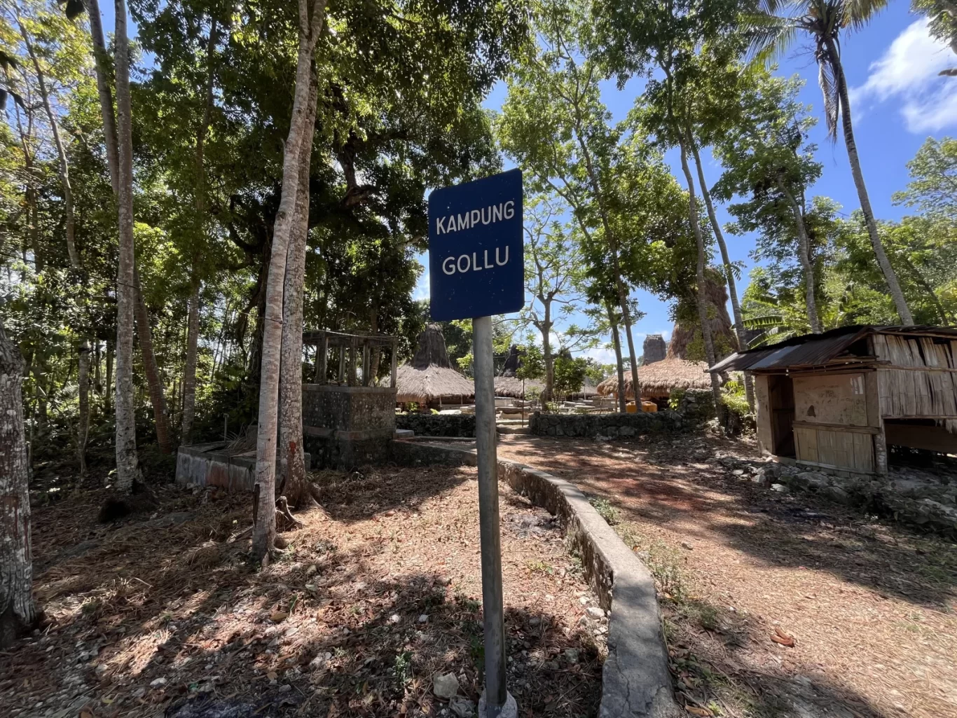 A blue sign for "Kampung Gollu" stands on a dirt path leading into a traditional village with thatched-roof houses and tall green trees under a bright sky.