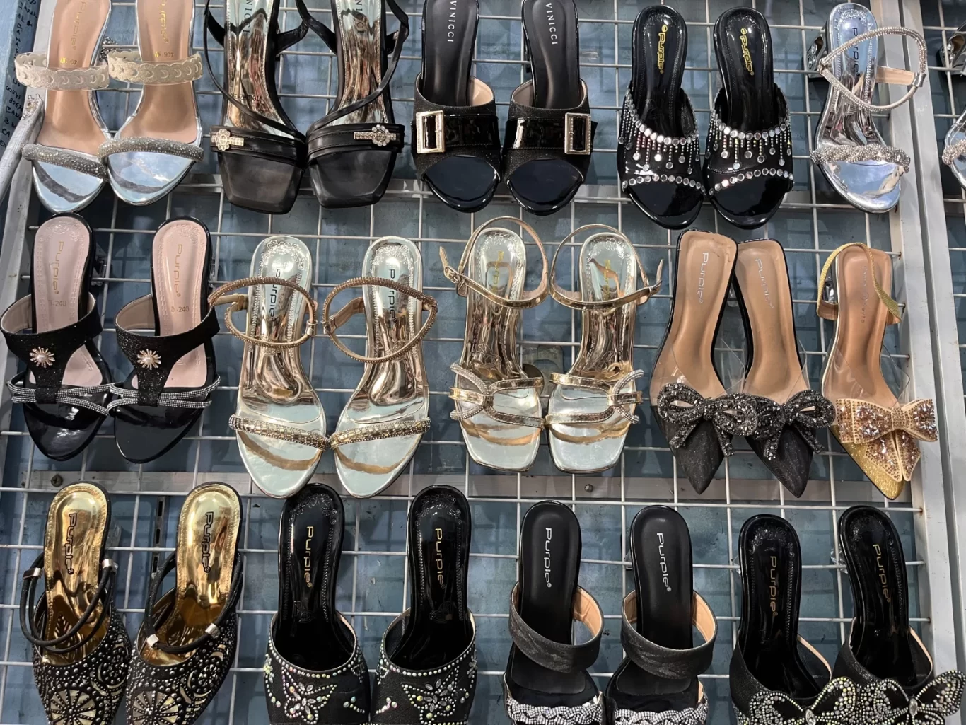Numerous pairs of women's high heels and sandals, adorned with glitter and rhinestones, are neatly displayed on a metal grid rack in a store.