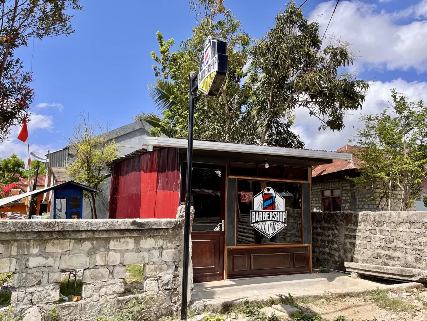 The dark brown exterior of Kilducy Barbershop stands behind a grey stone wall under a bright blue sky in Loli, Sumba.