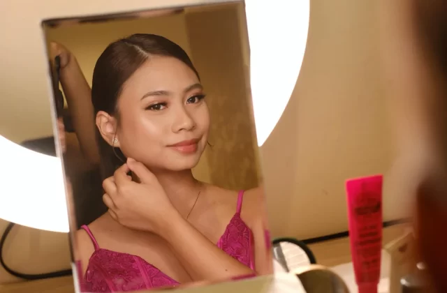 A young woman with subtle makeup and dark hair pulled back looks into a mirror, touching her ear, wearing a fuchsia pink lace top.