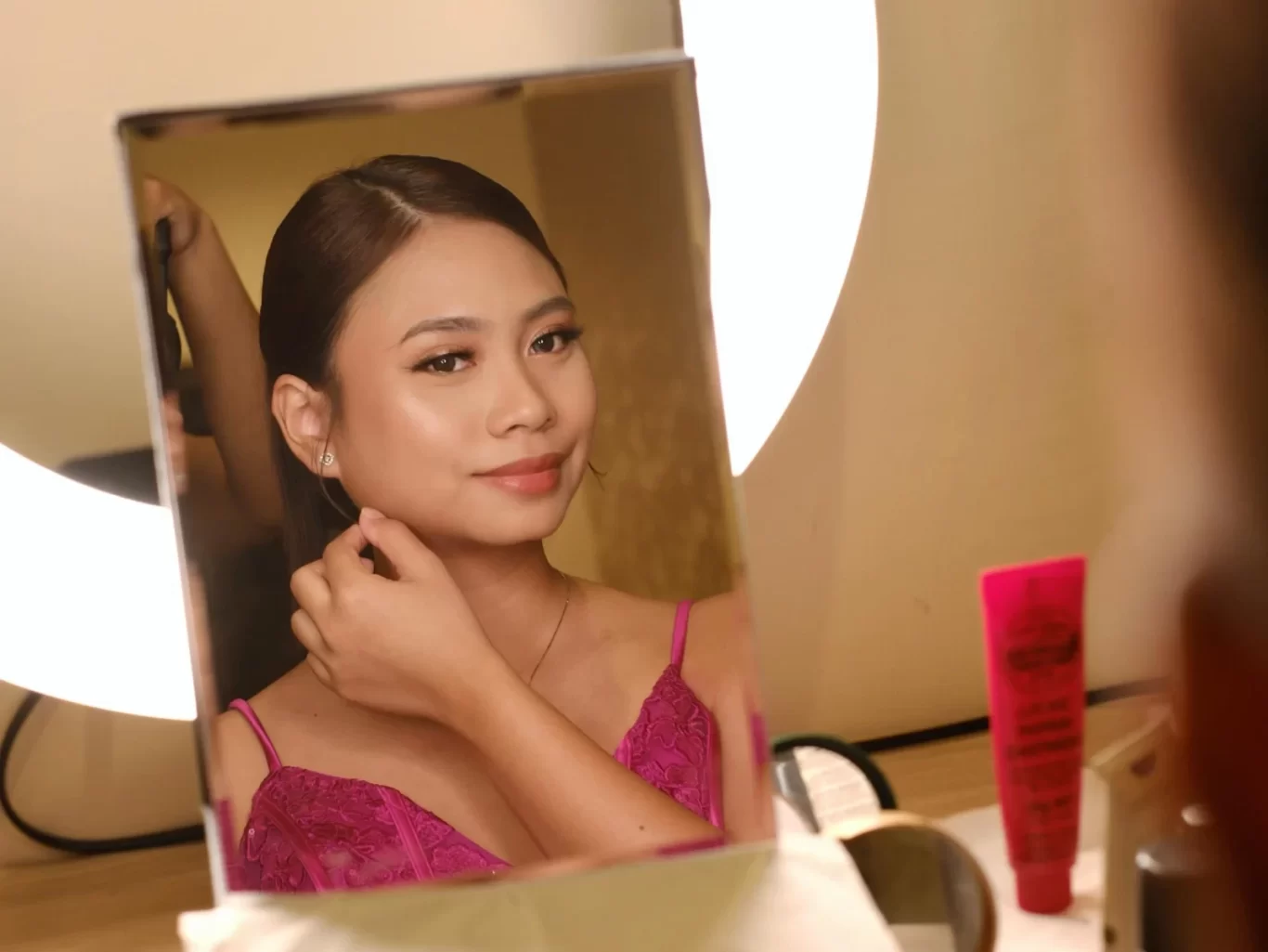 A young woman with subtle makeup and dark hair pulled back looks into a mirror, touching her ear, wearing a fuchsia pink lace top.