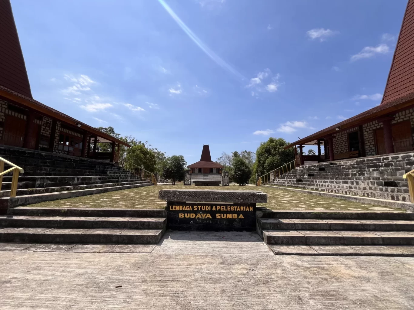 Two traditional Sumba buildings with high red roofs flank a central path leading to a smaller building under a bright blue sky, with a sign for 'Budaya Sumba' in the foreground.