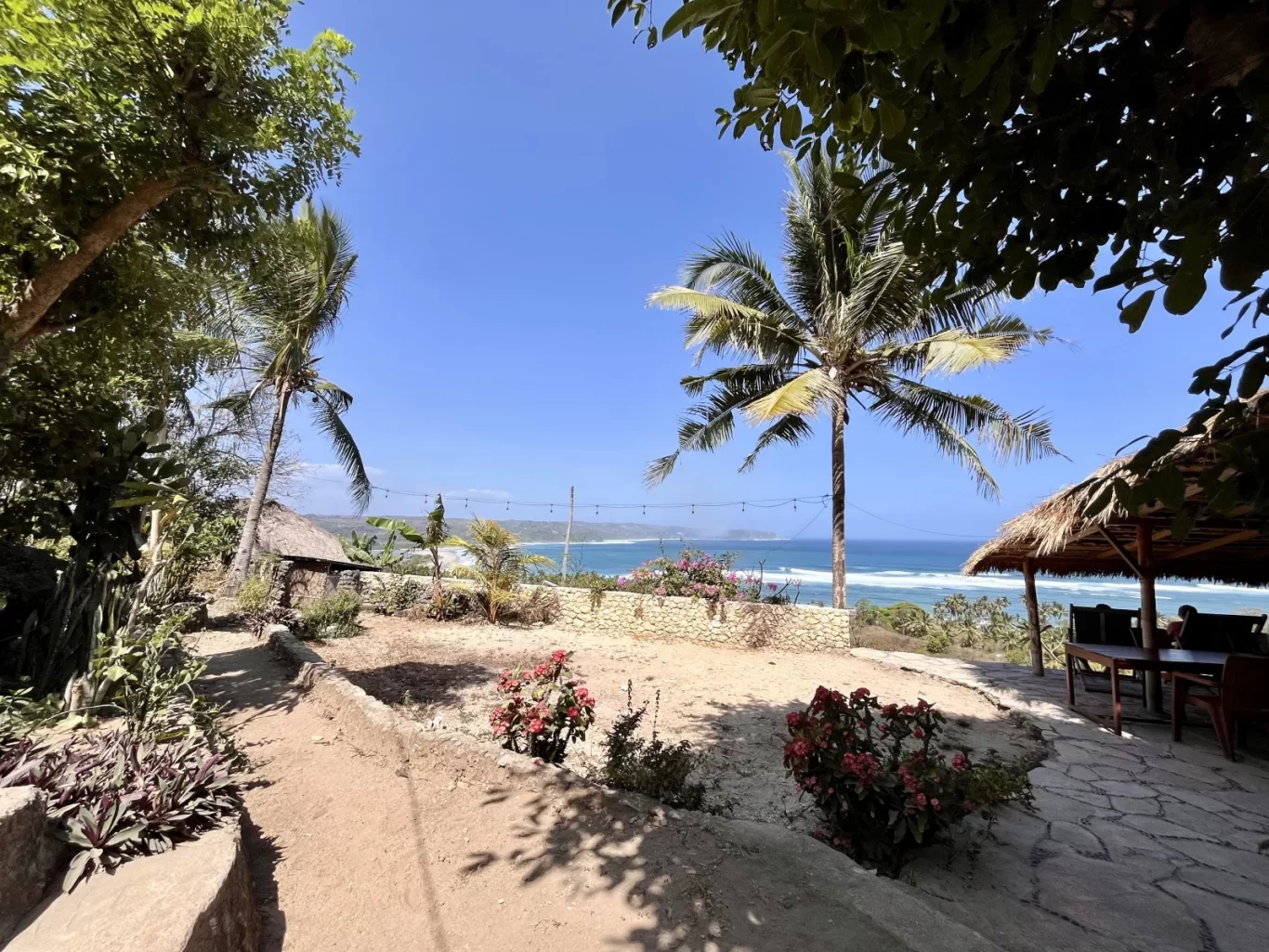 A sunny view from a Sumba hill shows two palm trees, a thatched-roof structure, and flowering plants overlooking a blue ocean with waves.