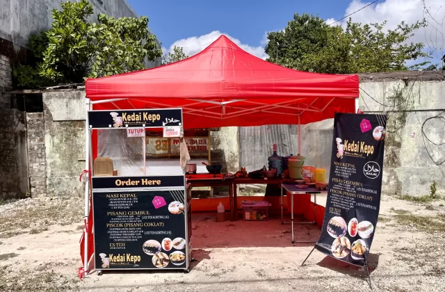 A red tent food stall named "Kedai Kepo" with black and white menus stands on a dirt patch against a concrete wall and green trees under a blue sky.